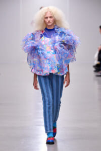Model walking a runway in a layered blue and pastel ruffled top over a printed dress, paired with blue striped jeans and colorful socks and shoes.