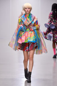 Model on a runway wearing a bright, sheer, multicolored oversized coat over a striped skirt and black ankle boots.