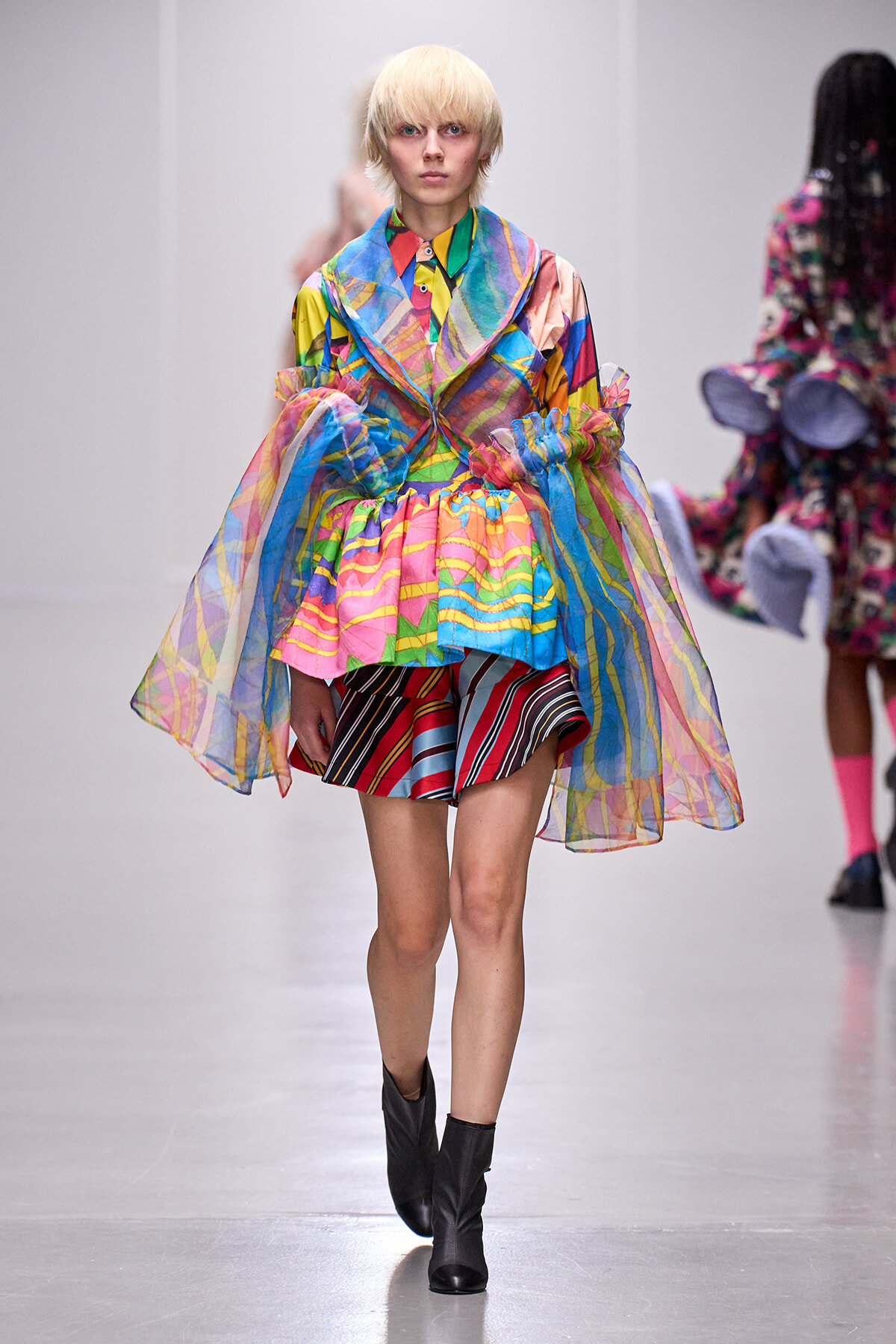 Model on a runway wearing a bright, sheer, multicolored oversized coat over a striped skirt and black ankle boots.