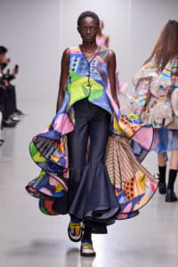 Model walks the runway in a vibrant, abstract patchwork dress with a high-low, voluminous hem over dark wide-legged pants; bold colors and shapes dominate the design.