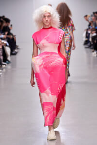 Model walks the runway in a hot-pink abstract-print sleeveless dress/jumpsuit with a white abstract pattern, paired with light-colored sneakers; audience lines the runway in the background.