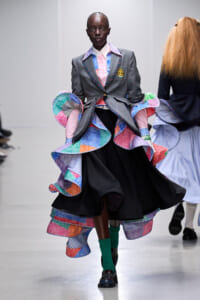 Model walking the runway in a gray blazer with crest, paired with a voluminous multicolor ruffled dress and bright green socks.