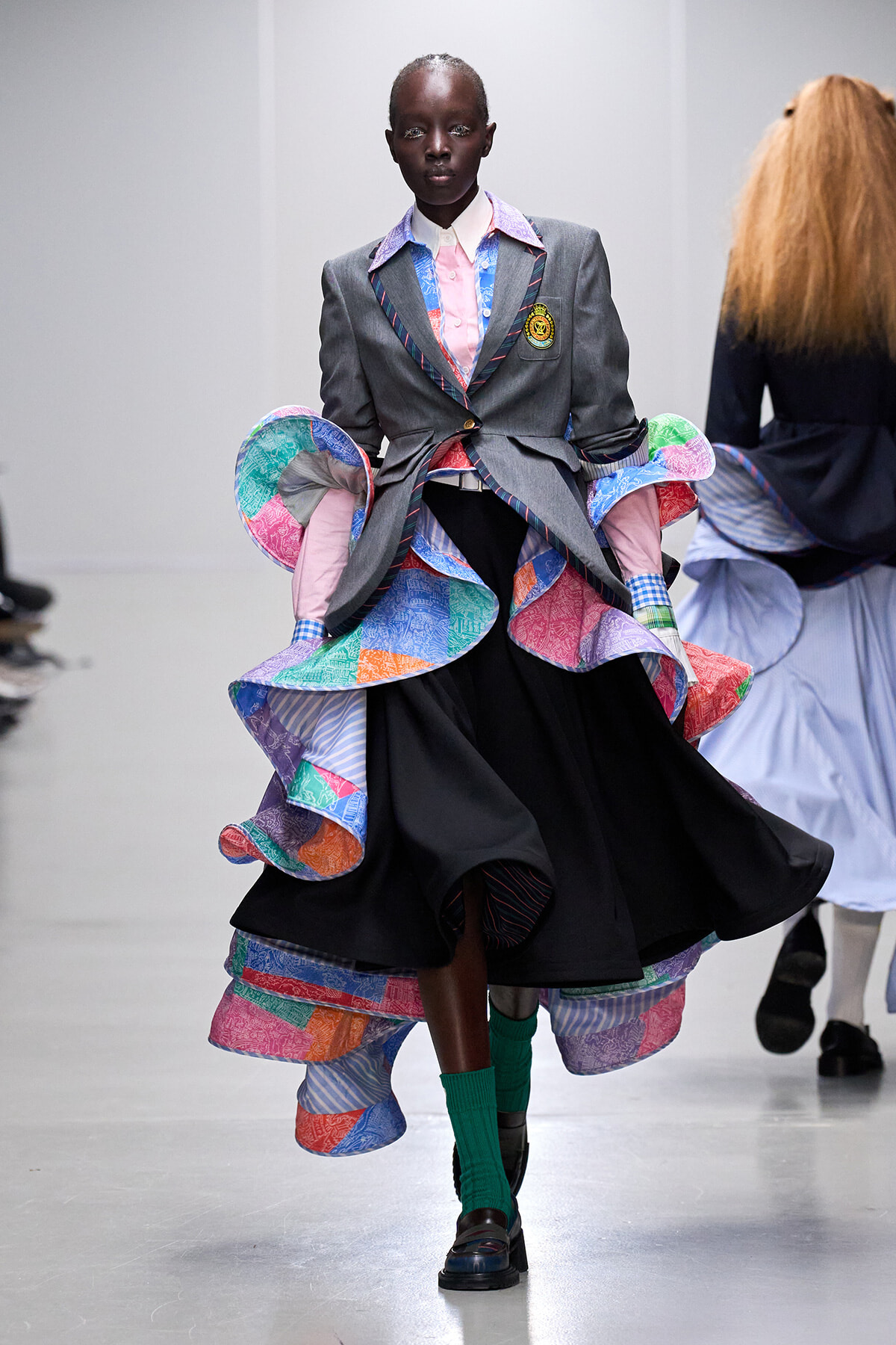 Model walking the runway in a gray blazer with crest, paired with a voluminous multicolor ruffled dress and bright green socks.