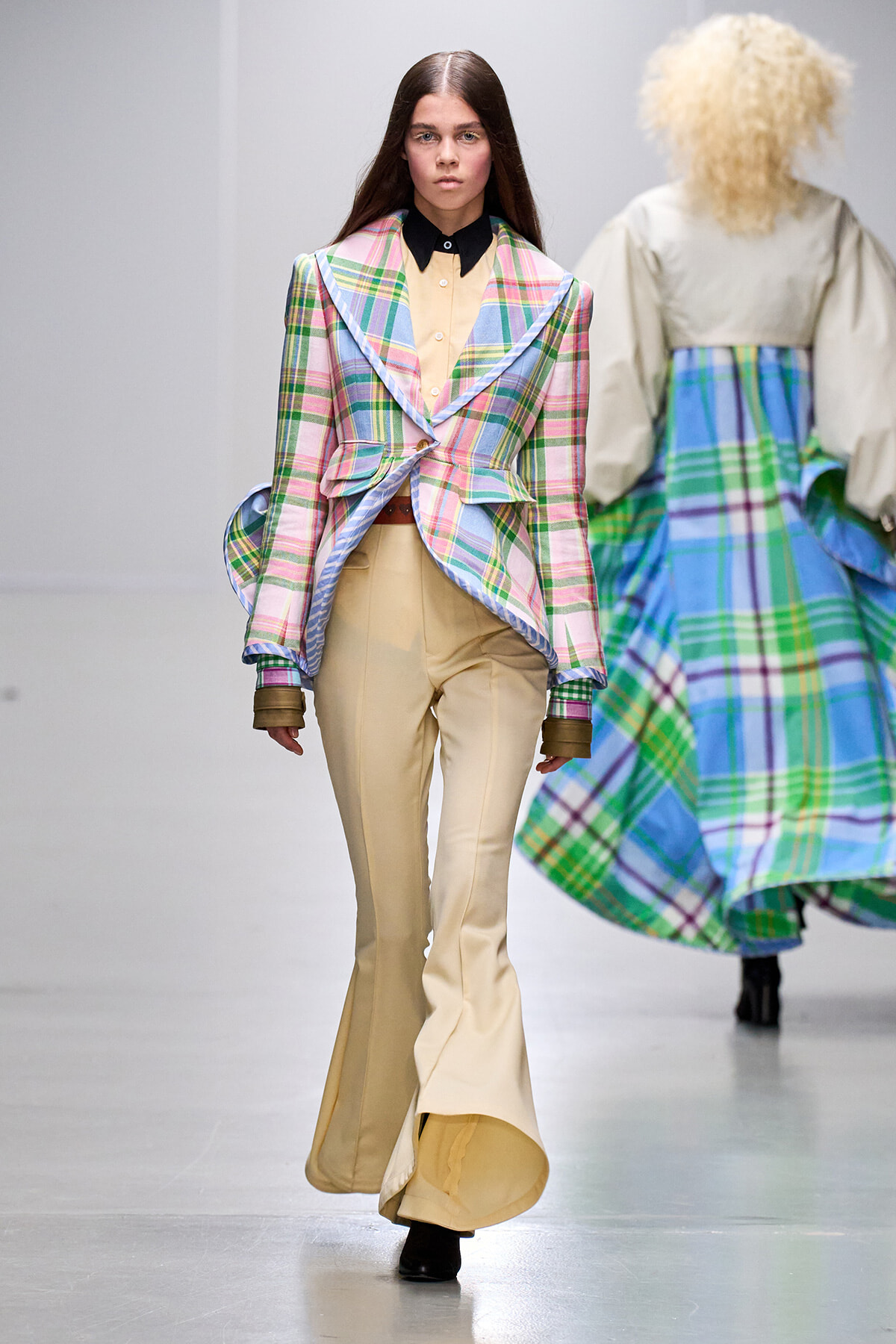Female model on a runway wearing a pastel plaid blazer over beige wide-leg pants and a black-collared shirt.