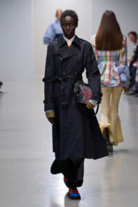 Model on a runway wearing a long black trench coat with a belt, carrying a patterned crossbody bag, and wearing bright socks.
