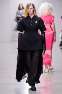 Model walking a fashion show in a structured black outfit: short-sleeve top with gold emblem and long sheer skirt, black gloves, blonde hair.
