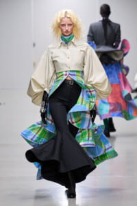 Model on a runway wearing a beige cropped blouse with voluminous sleeves and a colorful plaid skirt overlay over black pinstripe pants in motion.
