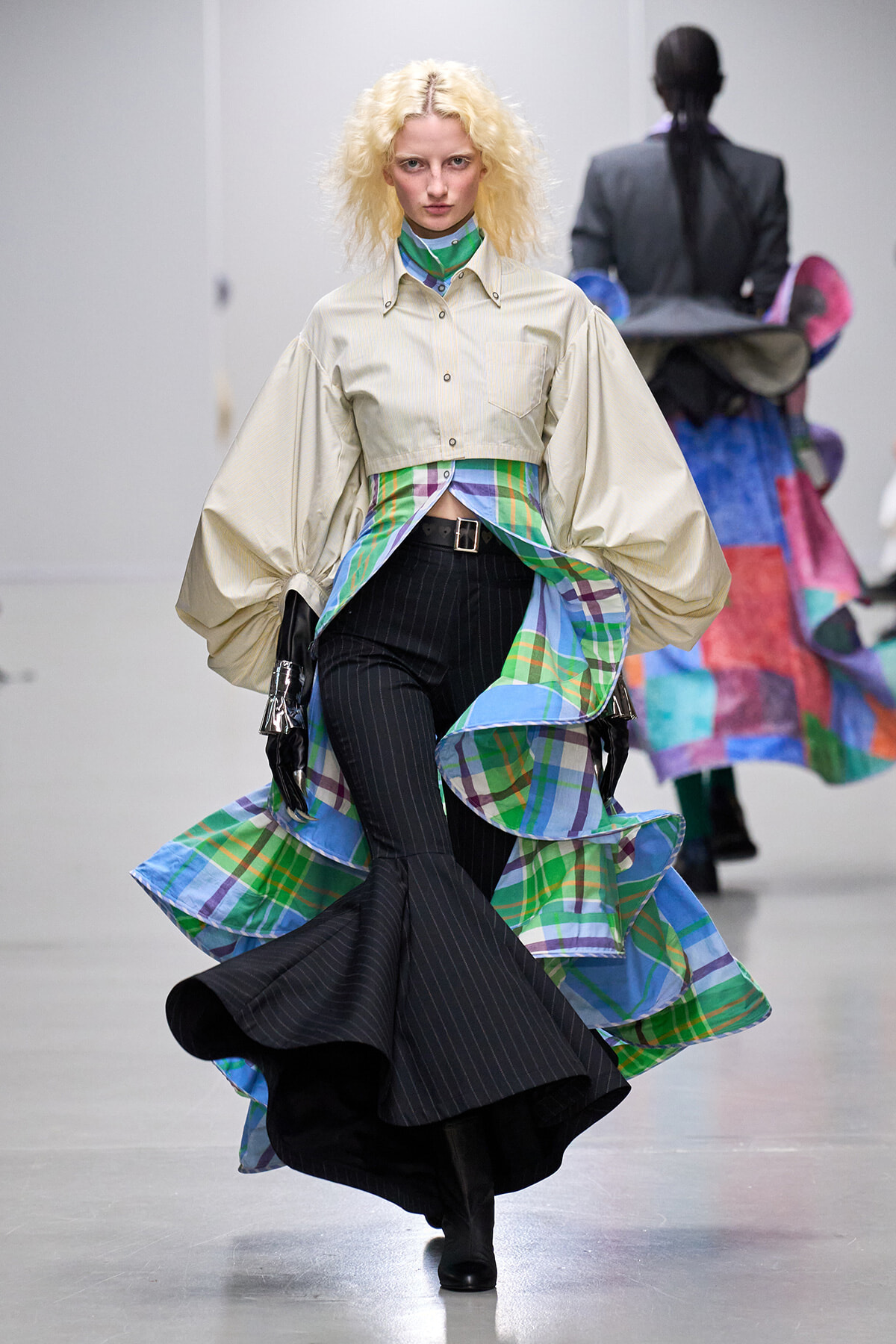 Model on a runway wearing a beige cropped blouse with voluminous sleeves and a colorful plaid skirt overlay over black pinstripe pants in motion.