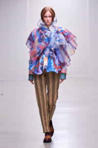 Model on a runway wearing a voluminous sheer multicolored ruffled top with brown vertical striped pants.