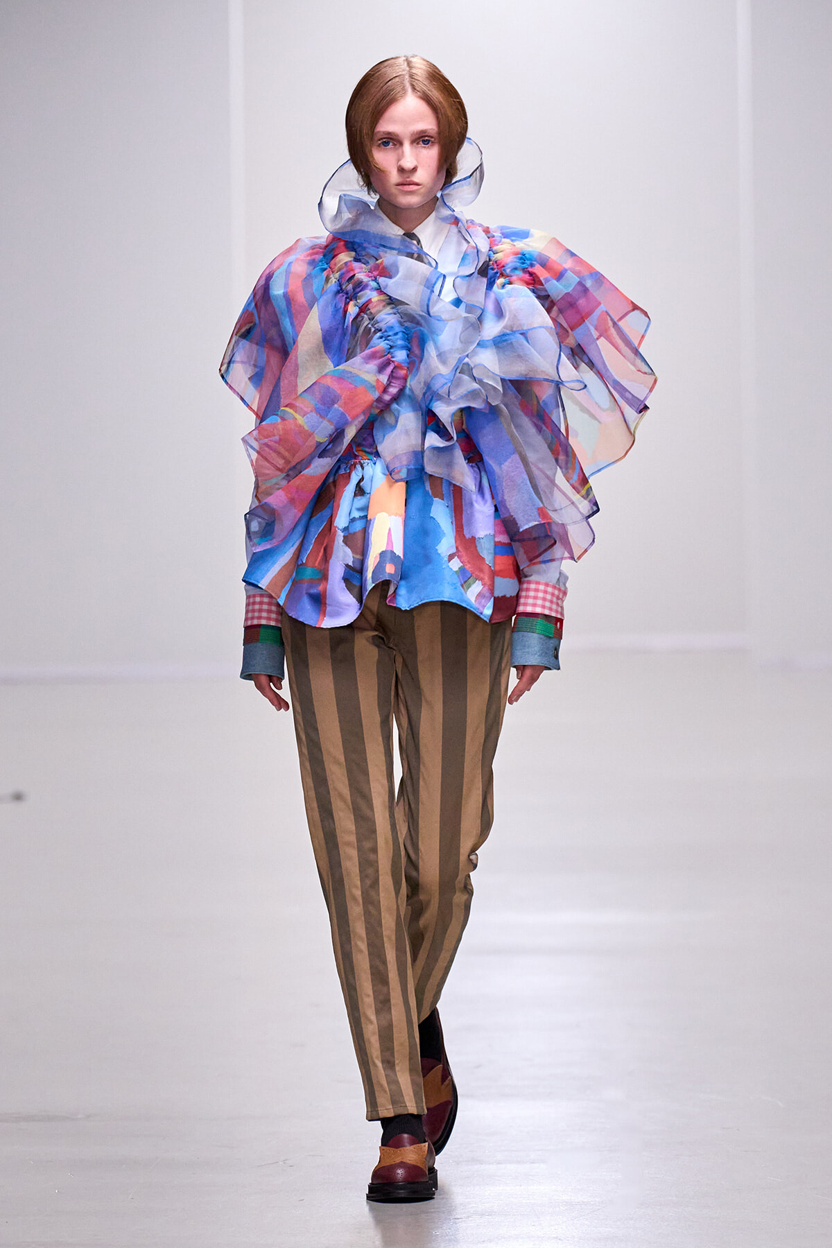 Model on a runway wearing a voluminous sheer multicolored ruffled top with brown vertical striped pants.