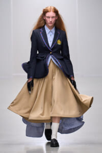 Model on a runway wearing a navy blazer with crest, striped shirt, and a voluminous beige skirt, with black gloves.
