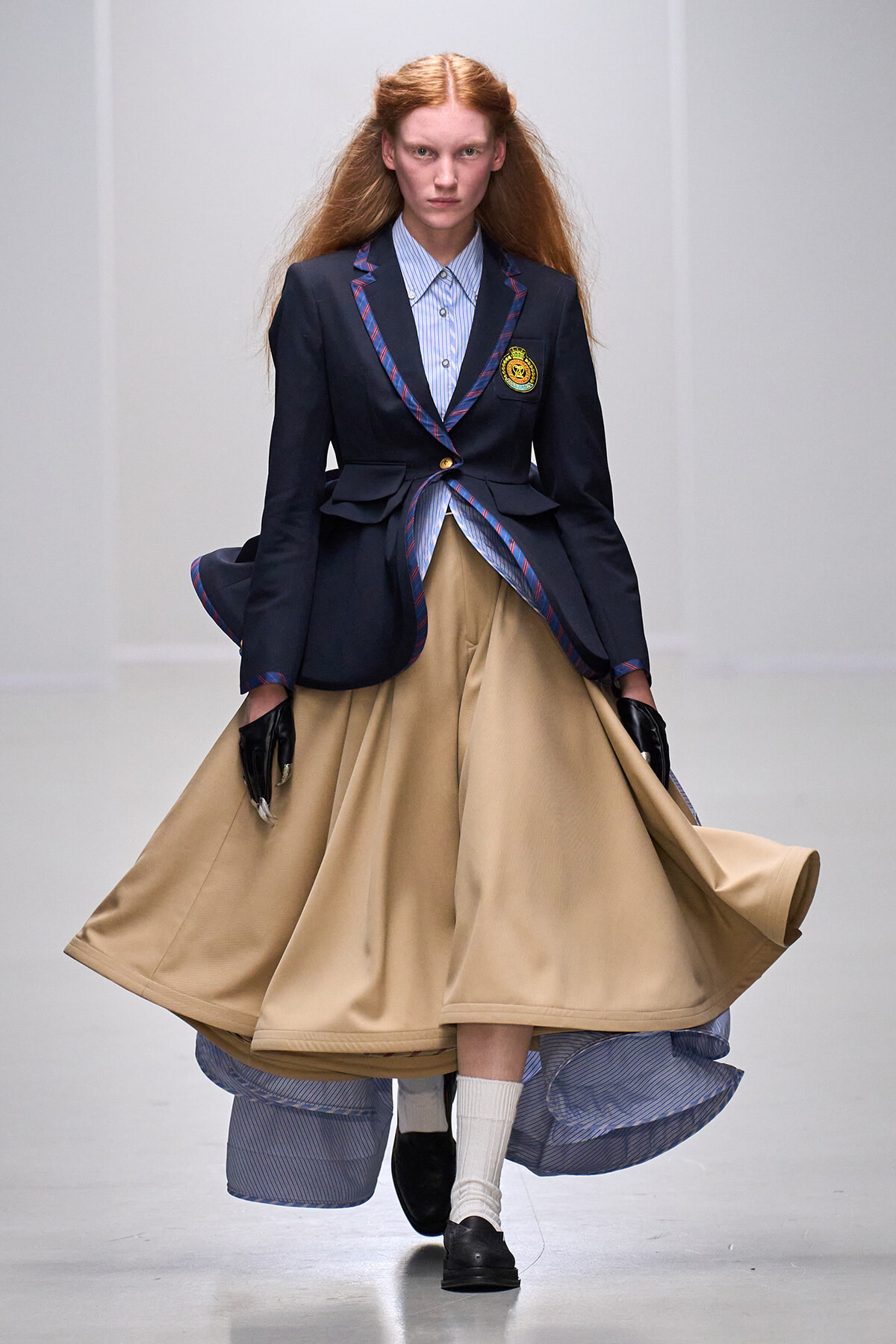 Model on a runway wearing a navy blazer with crest, striped shirt, and a voluminous beige skirt, with black gloves.