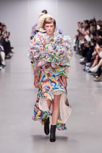 Model walks the runway in a voluminous, multicolored ruffled gown with bold layered fabric details.