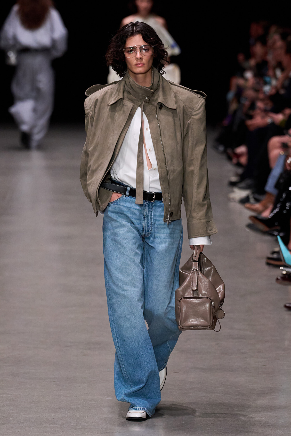 Model walking a runway in an oversized taupe jacket over a white shirt, light blue wide-leg jeans, black belt, and taupe handbag with sunglasses.