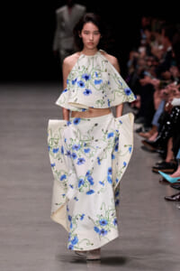 Model on a runway wearing a white two-piece gown with blue flowers and green vines, audience in the background.