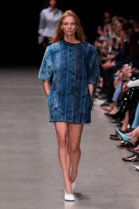 Model walks the runway in a knee-length denim striped shift dress with frayed hem and white flats, audience visible on the sides.