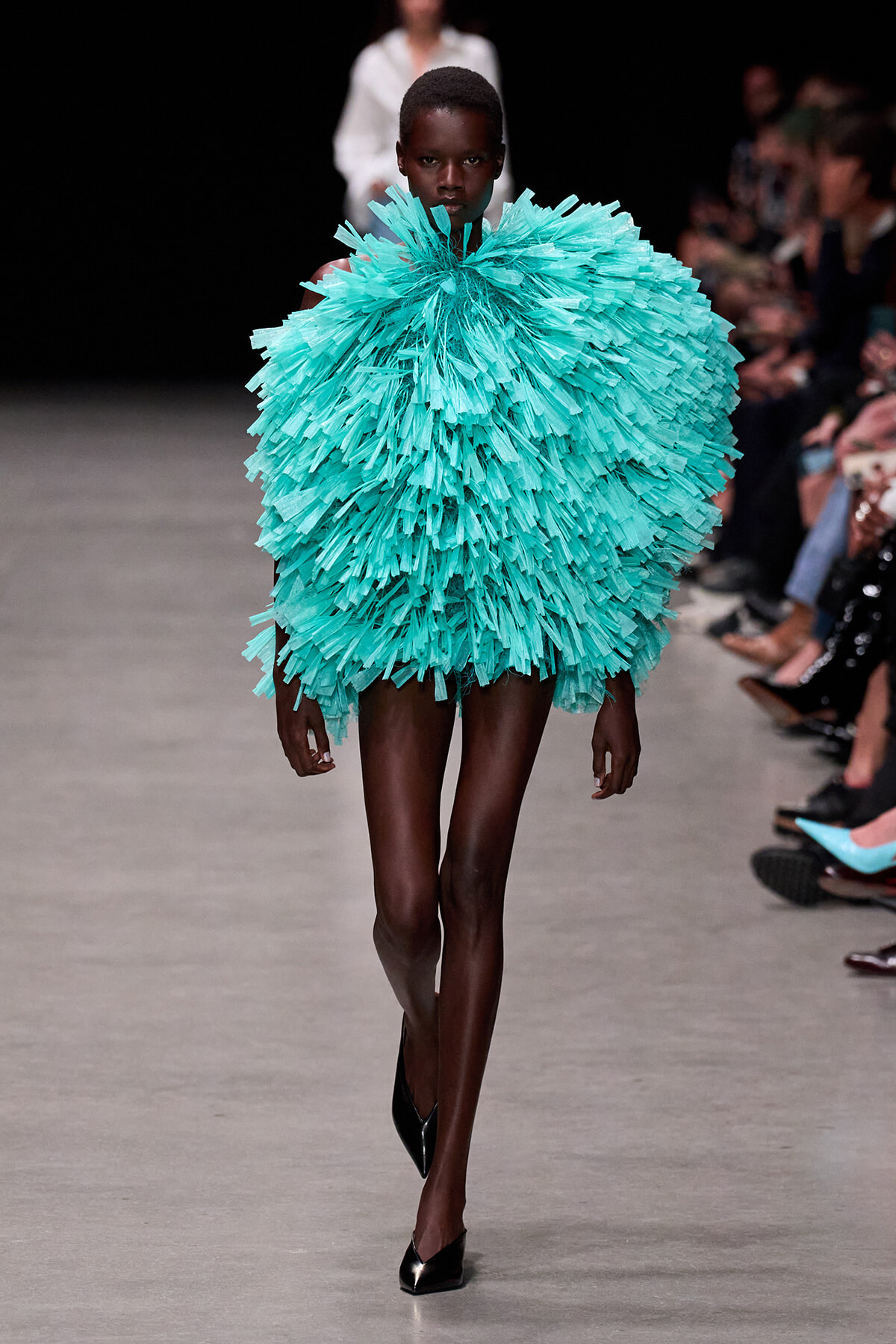 Model walks the runway in a voluminous turquoise fringe garment that envelops her torso, paired with black heels and a dark backdrop audience.