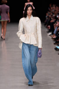 Model walking a runway in a cream textured button-up top and wide-leg blue jeans, holding a small purple clutch.