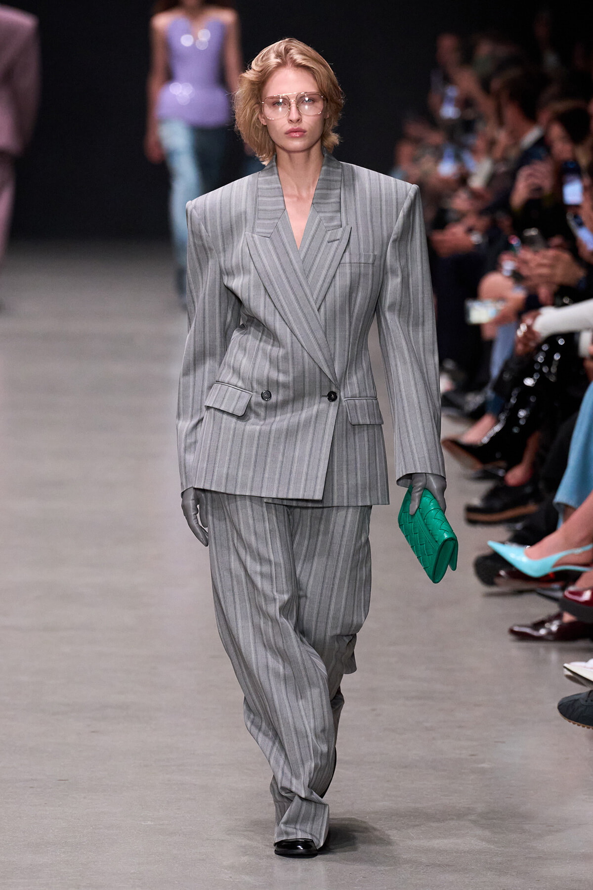 Model walks the runway in an oversized gray pinstripe double-breasted suit with broad shoulders, matching wide-leg pants, gray gloves, and a bright green clutch.