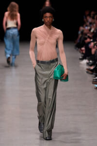 Model on a runway wearing a blush pink, twisted-draped top with gray wide-leg trousers, holding a bright green clutch.