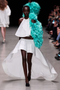 Model walks a runway in a white halter crop top and high‑low skirt, holding a turquoise feathered wrap over her shoulder.