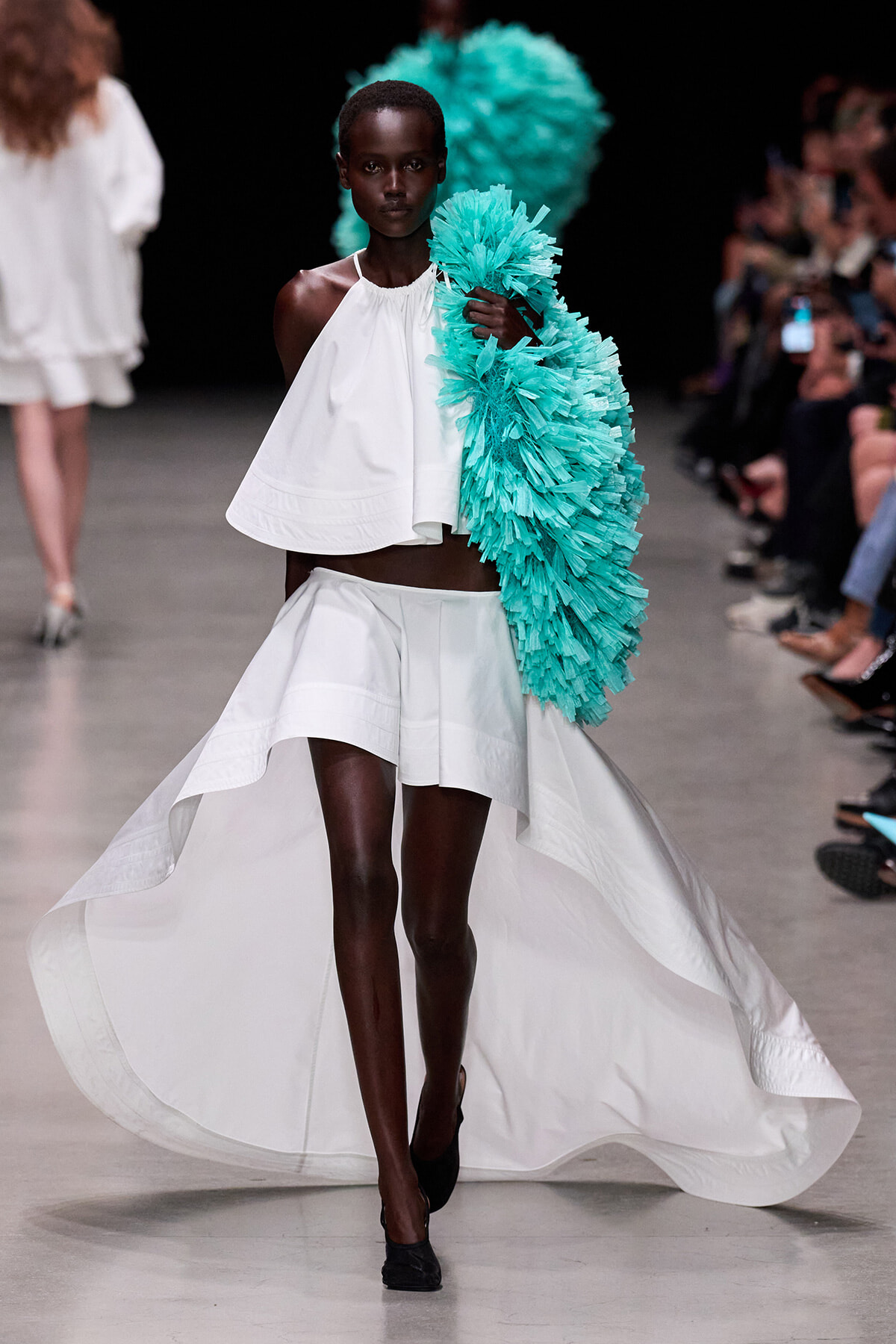 Model walks a runway in a white halter crop top and high‑low skirt, holding a turquoise feathered wrap over her shoulder.