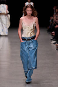 Model walking a runway in a cream satin lace camisole and oversized blue jeans with a black belt.