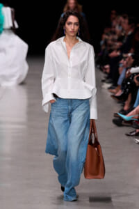 Model walks a fashion runway in a white button-up blouse tucked into oversized blue jeans, carrying a brown bag.