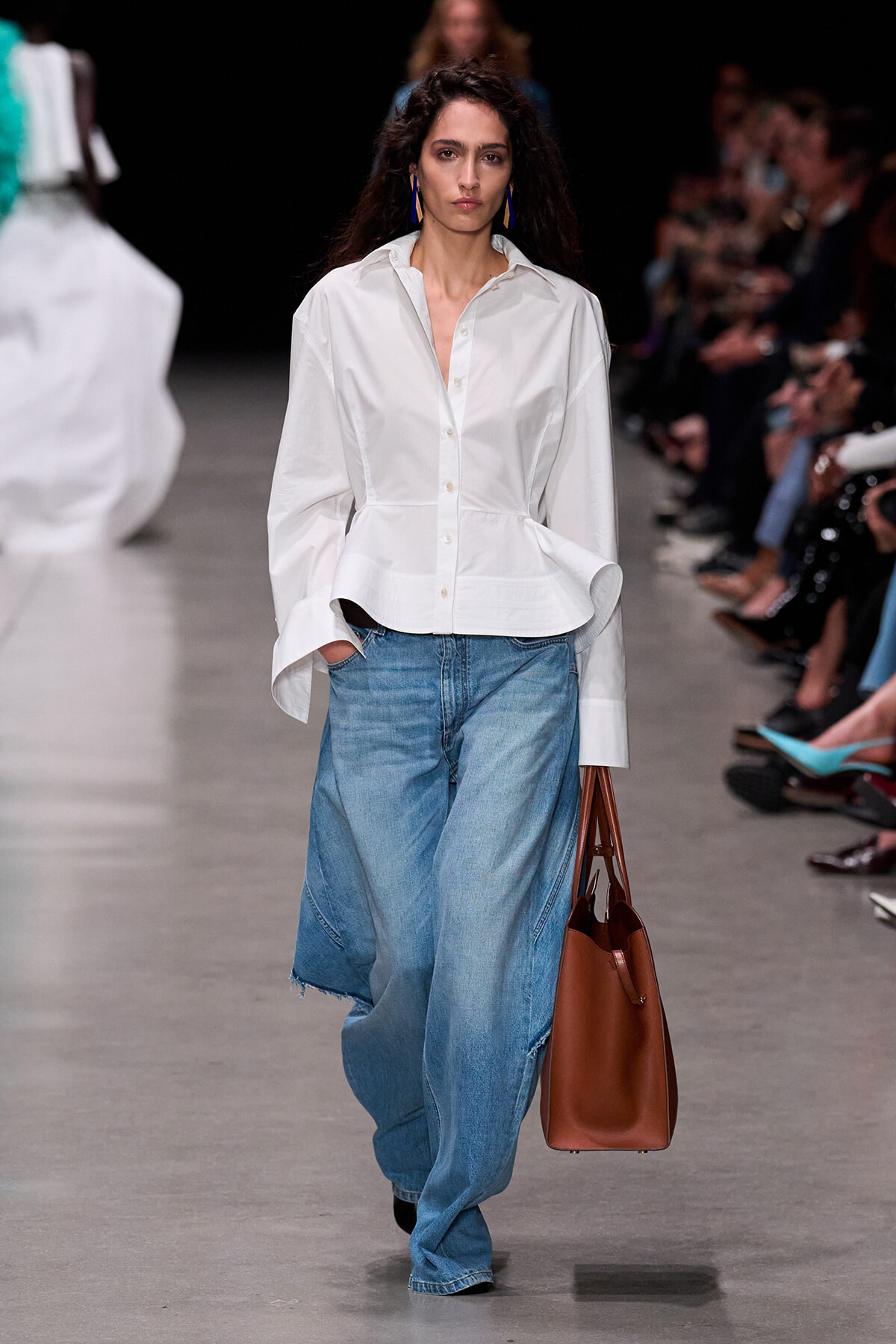 Model walks a fashion runway in a white button-up blouse tucked into oversized blue jeans, carrying a brown bag.