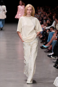 Female model walking a runway in a beige oversized T-shirt and sparkly cream sequined pants, audience watching.
