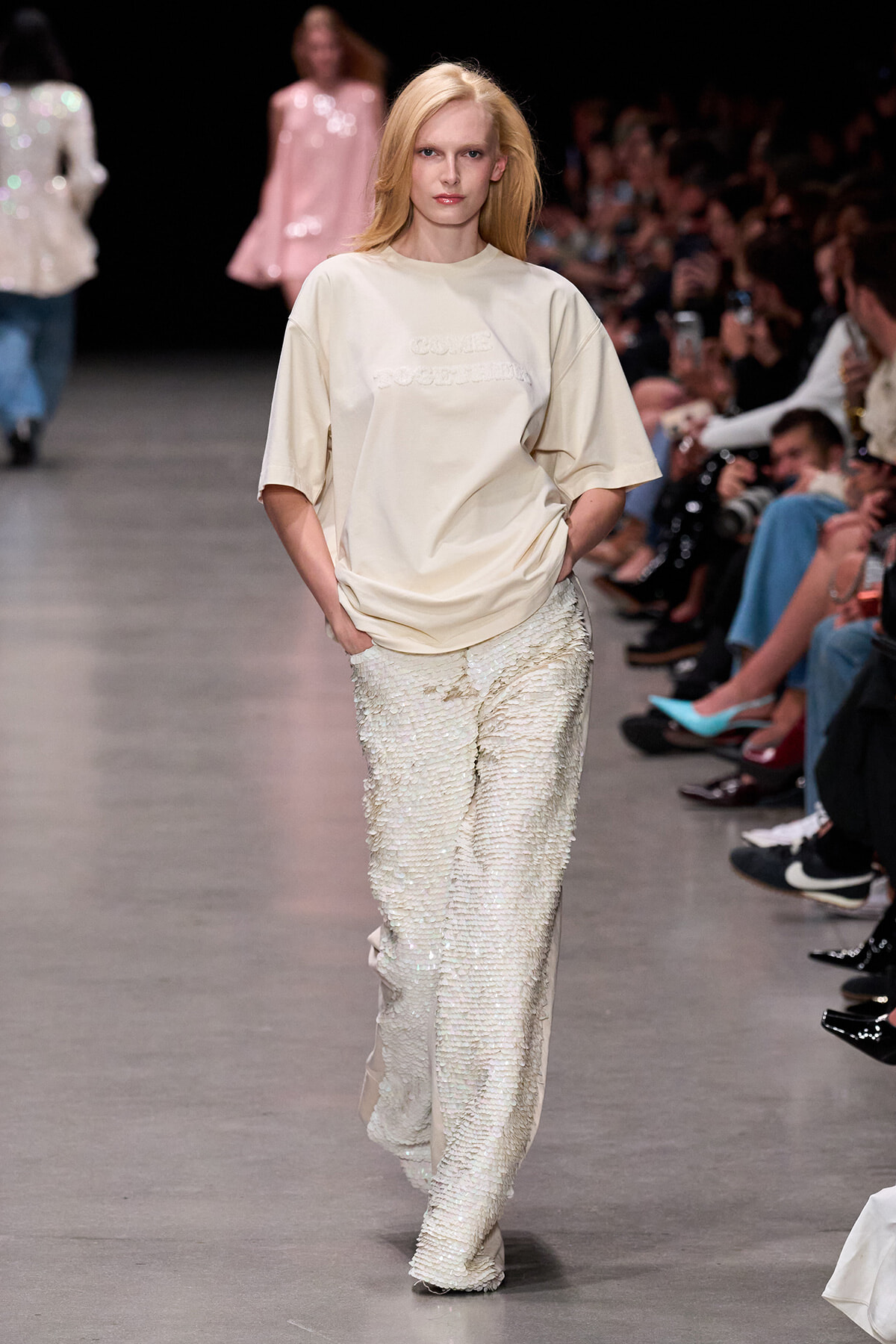 Female model walking a runway in a beige oversized T-shirt and sparkly cream sequined pants, audience watching.