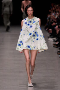 Model walking the runway in a sleeveless white mini dress with blue floral embroidery and green vines.