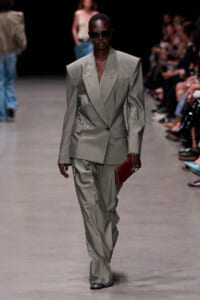 Model walks the runway in a light gray, oversized blazer with matching wide-leg pants and a red clutch.