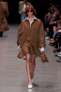 Model walking the runway in a loose brown button-up shirt worn over a striped shirt, holding a small brown quilted handbag and wearing white shoes with dark sunglasses.