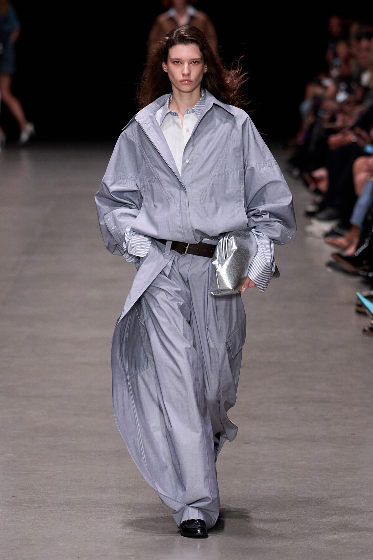 Model walks the runway in an oversized light-gray trench-style suit with a white shirt, cinched by a dark belt and paired with a metallic clutch.