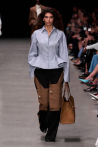 Runway model wearing a light blue structured peplum shirt and brown color-block cargo pants, carrying a brown handbag.