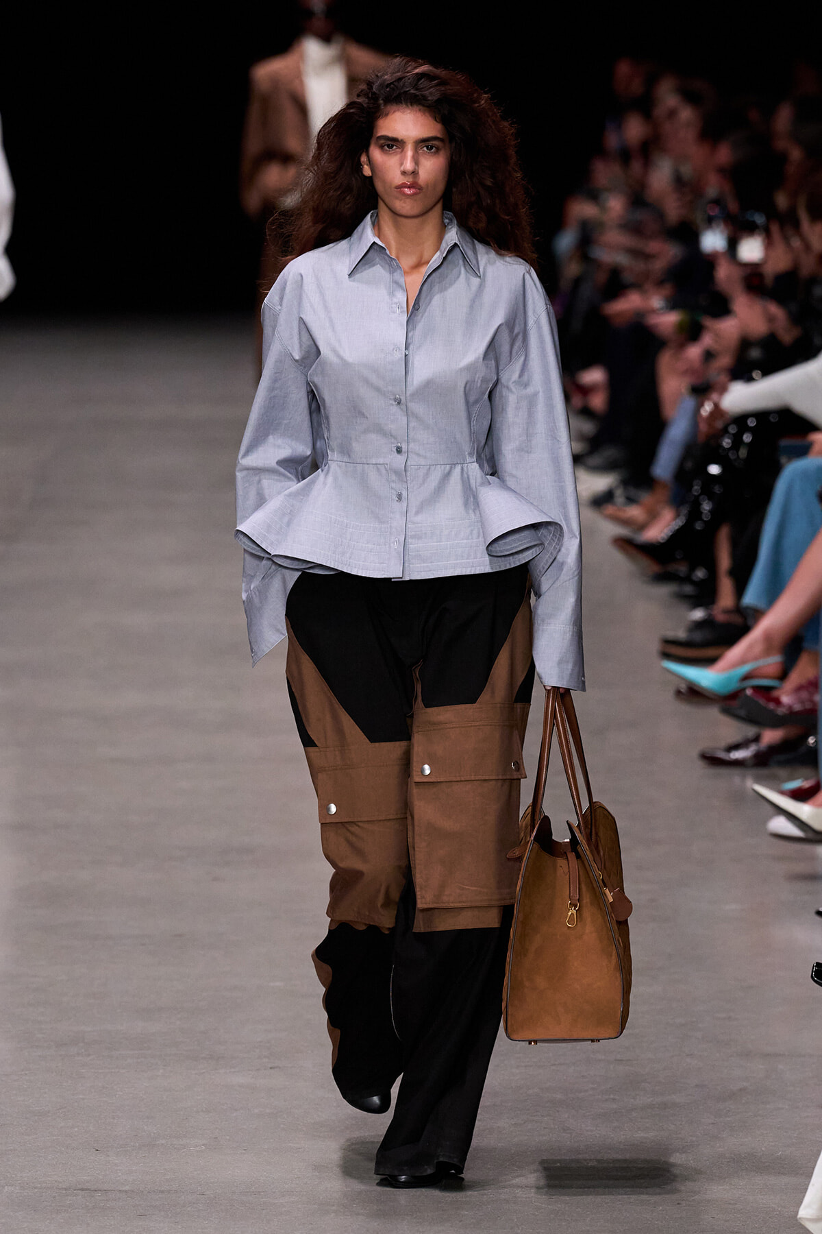 Runway model wearing a light blue structured peplum shirt and brown color-block cargo pants, carrying a brown handbag.