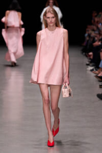 Model walks the runway in a pale pink sequined sleeveless shift dress, carrying a small pink handbag and red heels.