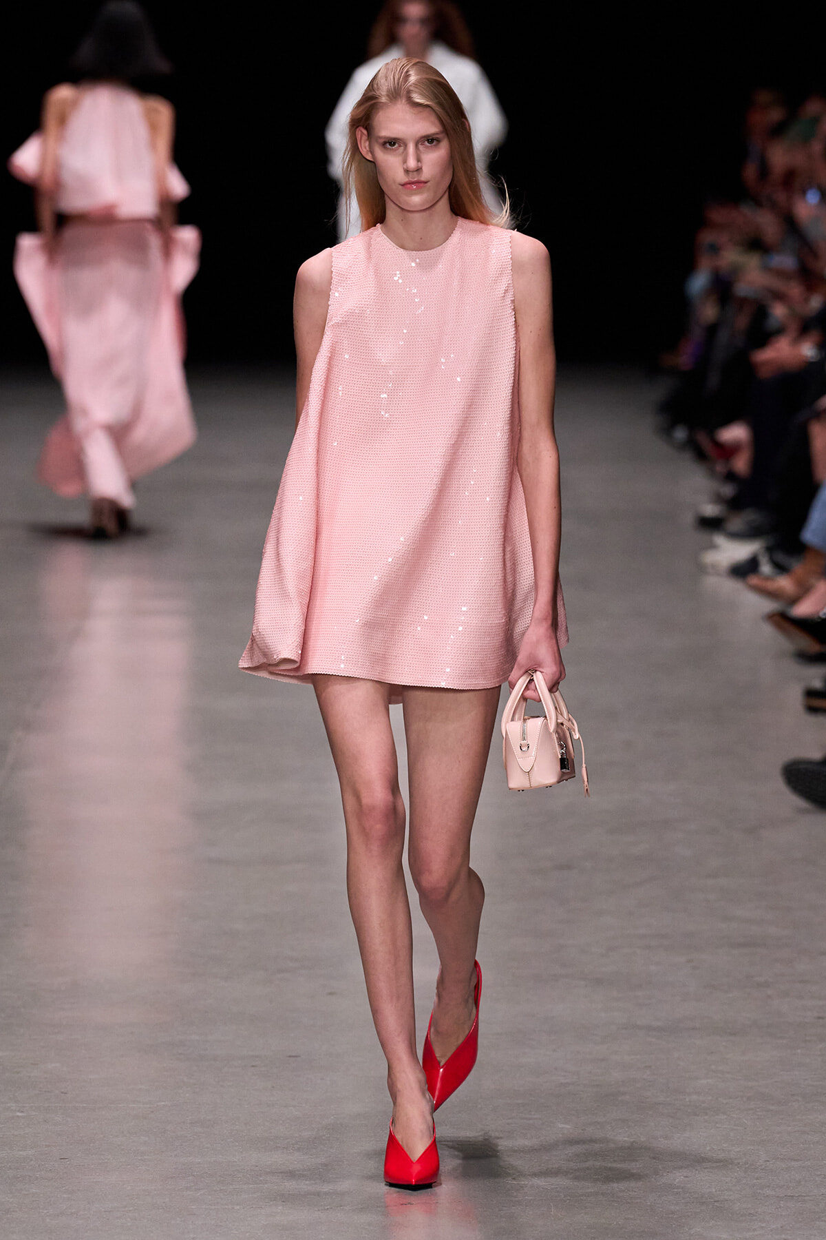 Model walks the runway in a pale pink sequined sleeveless shift dress, carrying a small pink handbag and red heels.