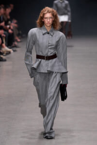 Female model walks the runway in a grey pinstriped suit with a wide dark belt, carrying a black clutch.