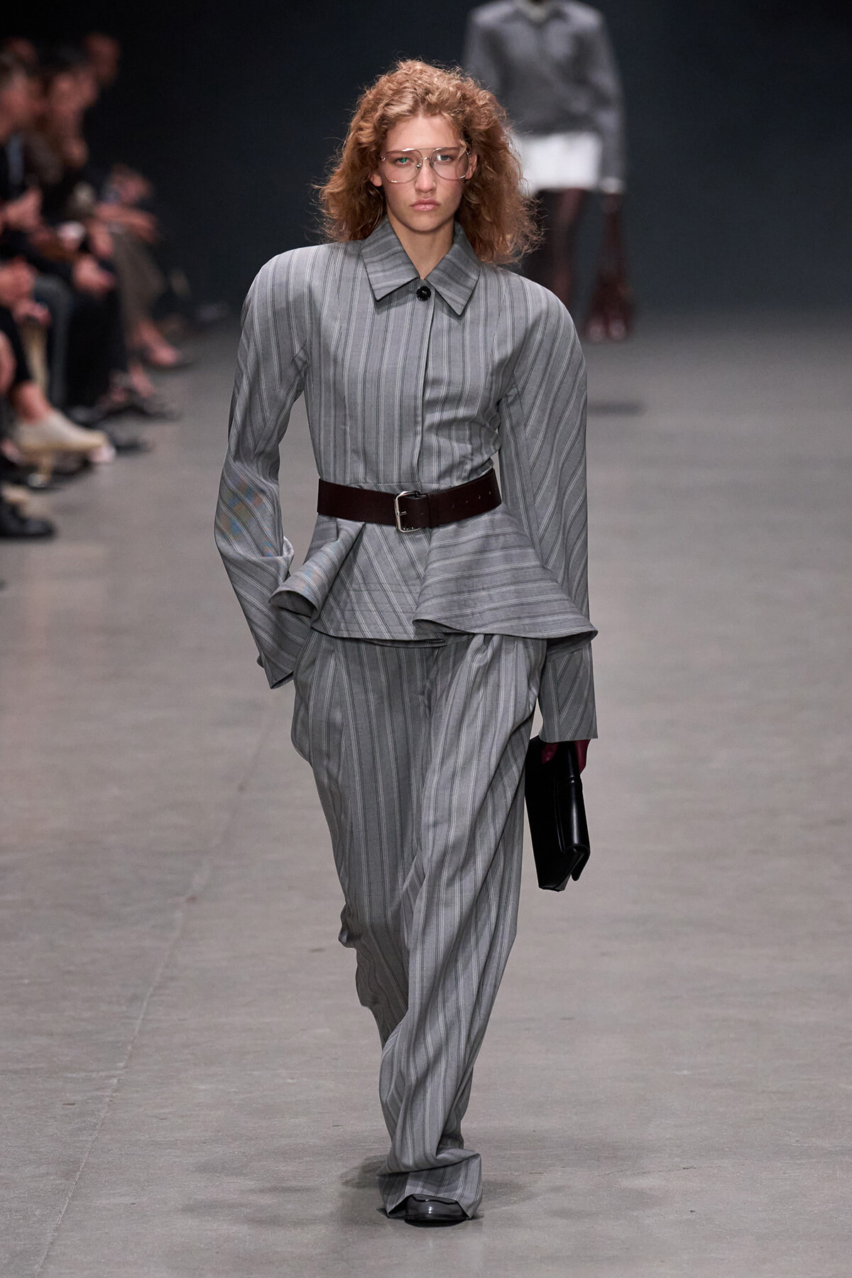 Female model walks the runway in a grey pinstriped suit with a wide dark belt, carrying a black clutch.