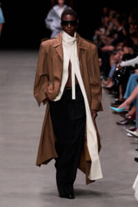 Model on a runway wearing an oversized tan trench coat over a white high-collar top and black wide-leg pants, with dark sunglasses.