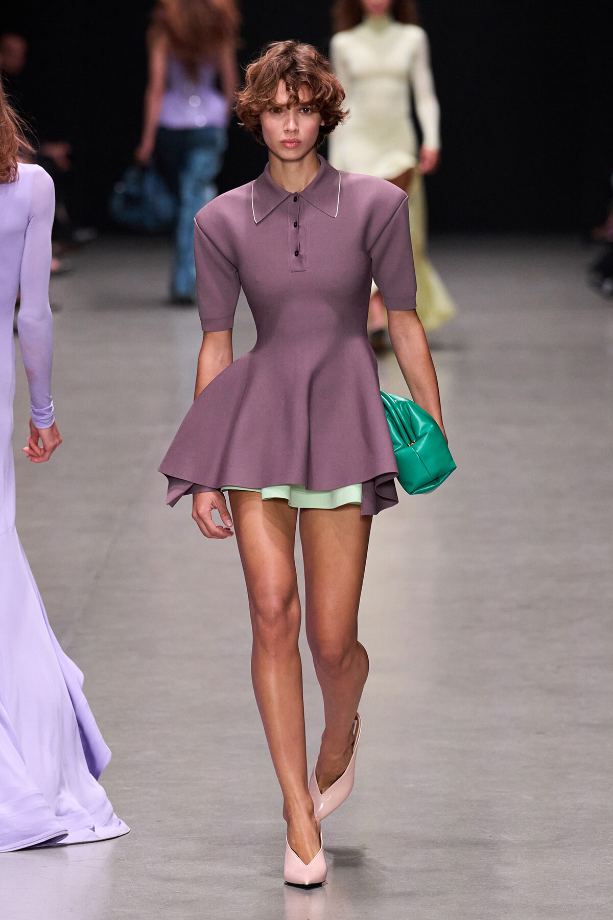Model walking a runway in a lavender peplum dress with short sleeves, mint green shorts peeking out, and pink pumps carrying a green clutch.
