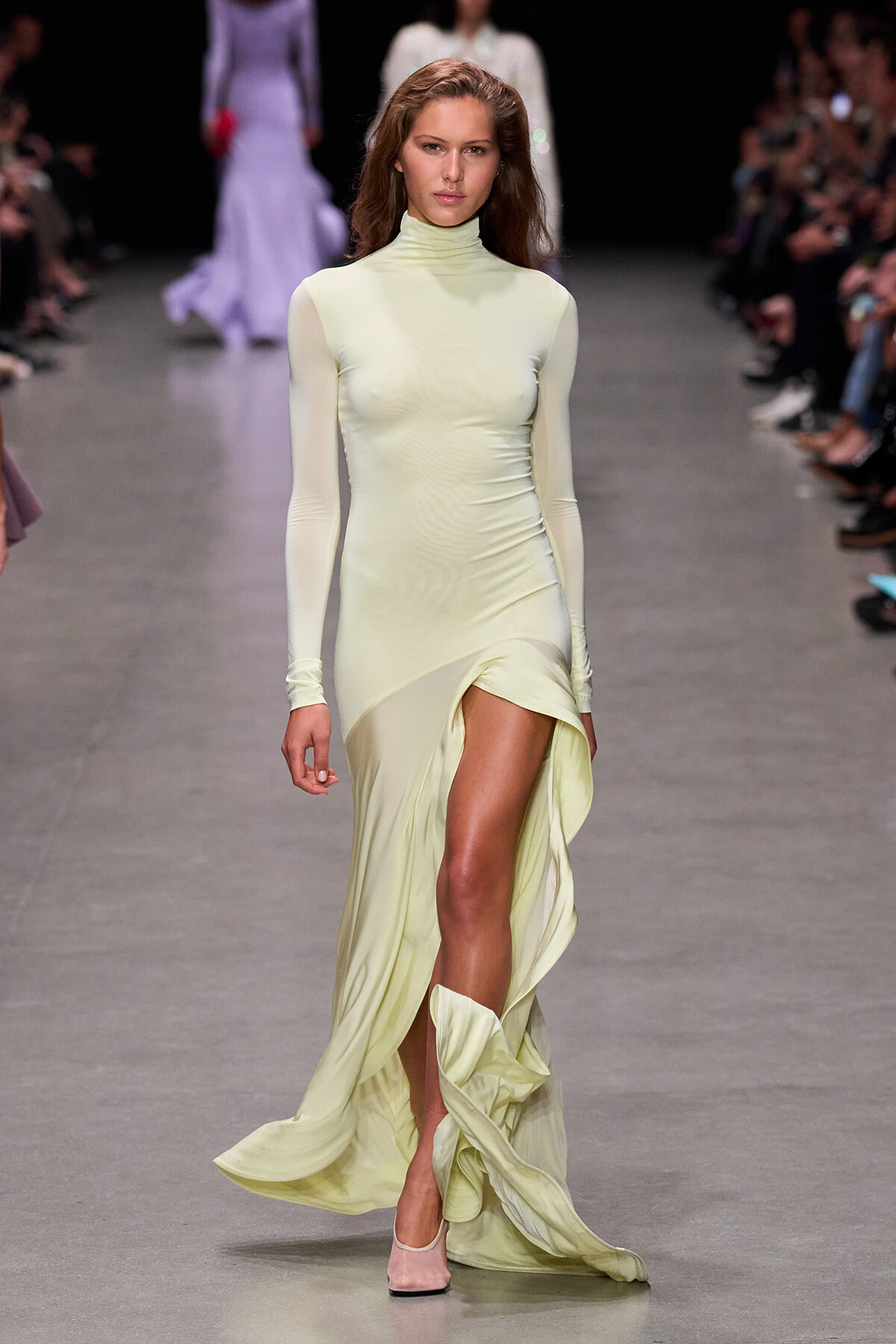 Model walks the runway in a pale lemon-yellow, high-neck, long-sleeve gown with a high slit revealing one leg, paired with pale pink heels.