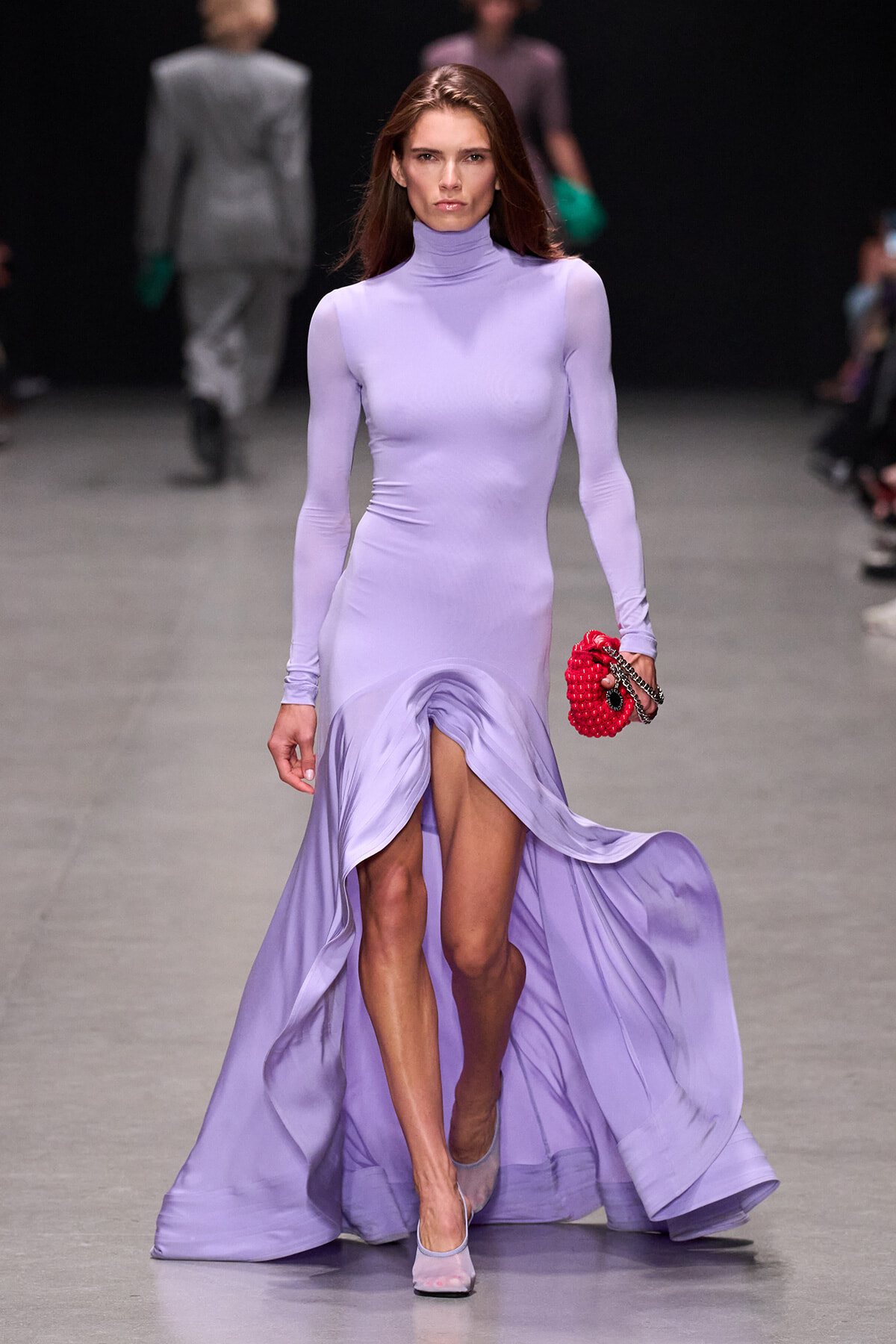 Model on a fashion runway wears a lavender turtleneck, long-sleeve gown with a high slit, carrying a bright red beaded clutch.