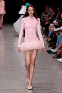 Model walking a runway in a pale pink, long-sleeve mini dress with a fluffy tulle skirt and matching pink heels.