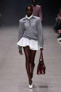 Model walks the runway in a gray long-sleeve polo over a white layered mini skirt, carrying a burgundy handbag.