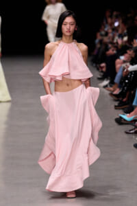Model walks the runway in a pale pink halter top with a draped cape-like overlay and a long flowing skirt.