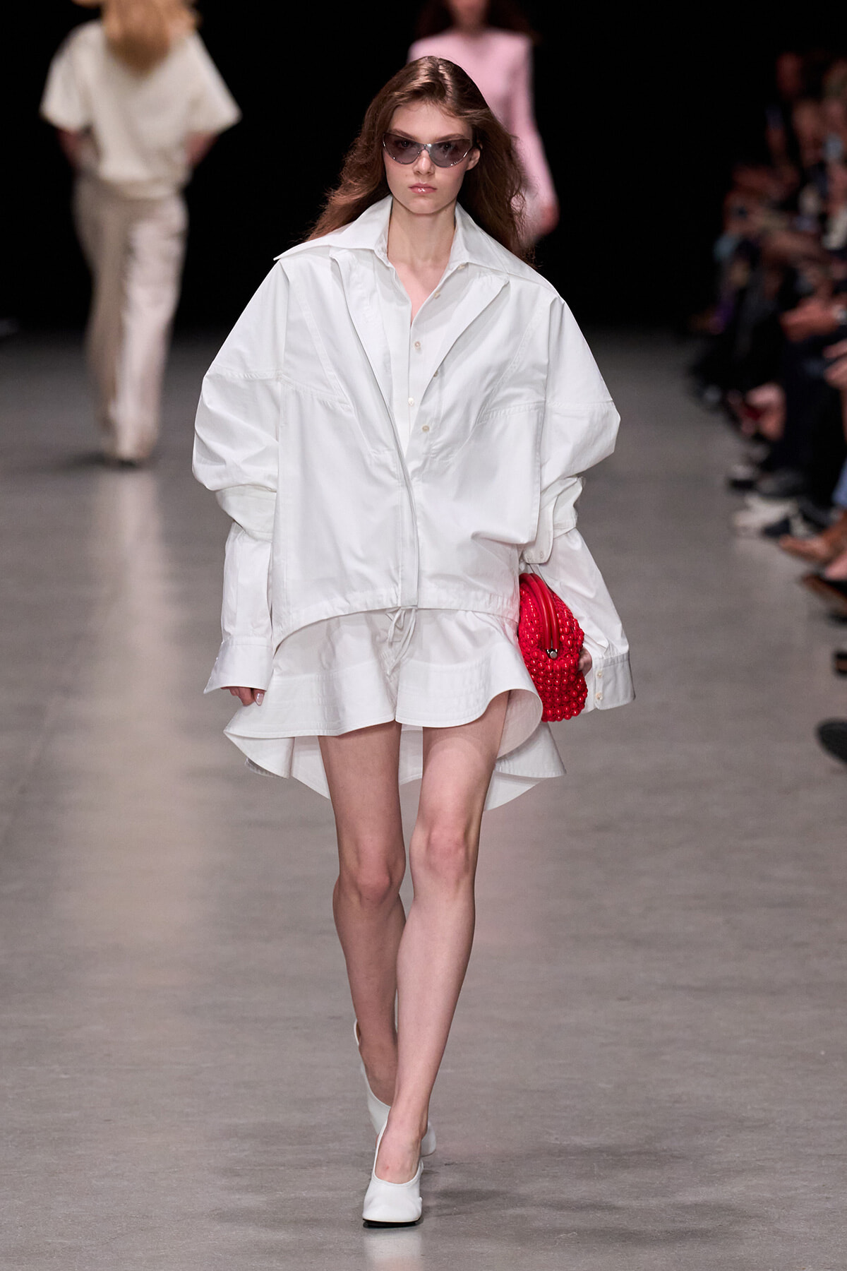 Model walking a runway in an oversized white shirt-jacket and short white skirt, carrying a red beaded clutch and wearing sunglasses on a fashion show runway.
