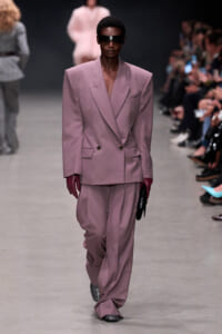 Model walks the runway in an oversized dusty-pink double-breasted suit with wide shoulders, burgundy gloves, sunglasses, and a black clutch.